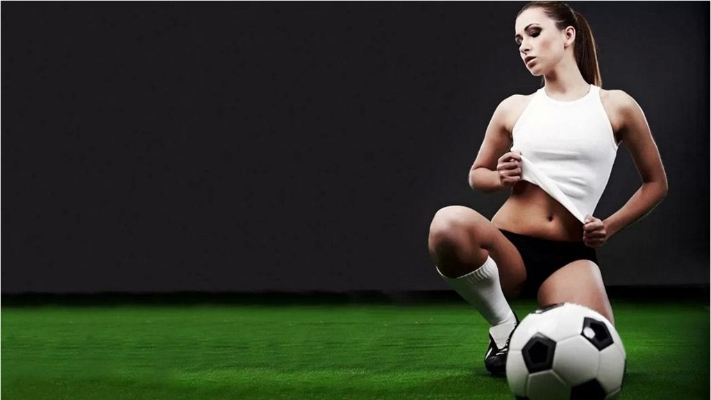 Девушка в черных трусах, белом топе, в гетрах и спортивной обуви сидит в откровенной позе на футбольном поле рядом с мячем
