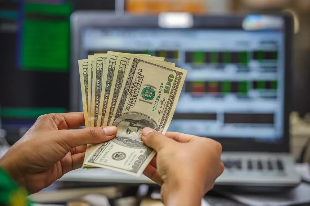 Веер из долларовых купюр разного номинала в руках человека на фоне открытого ноутбука