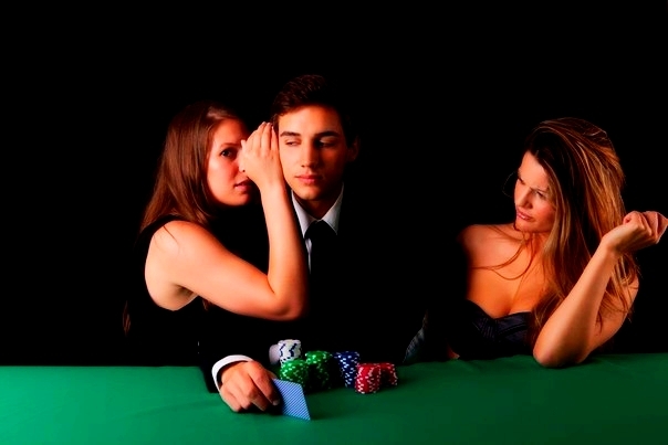 Парень за столом для покера с двумя красотками, одна из которых нашептывает ему на ухо