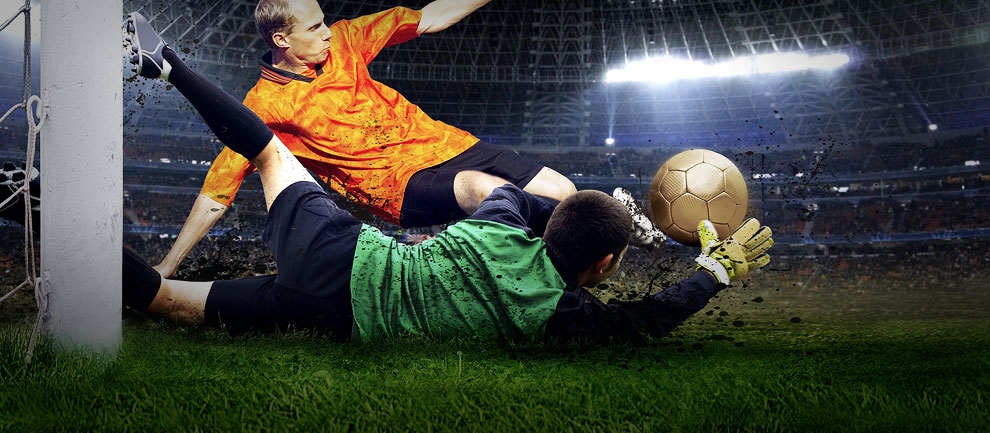 Футболист в зеленой футболке падает, забивая мяч в ворота, а вратарь в оранжевой футболке пытается поймать мяч