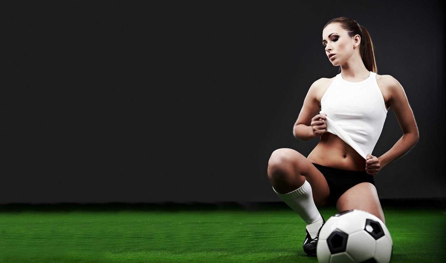 Спортсменка в черных трусах и белом топе позирует на футбольном поле в полумраке