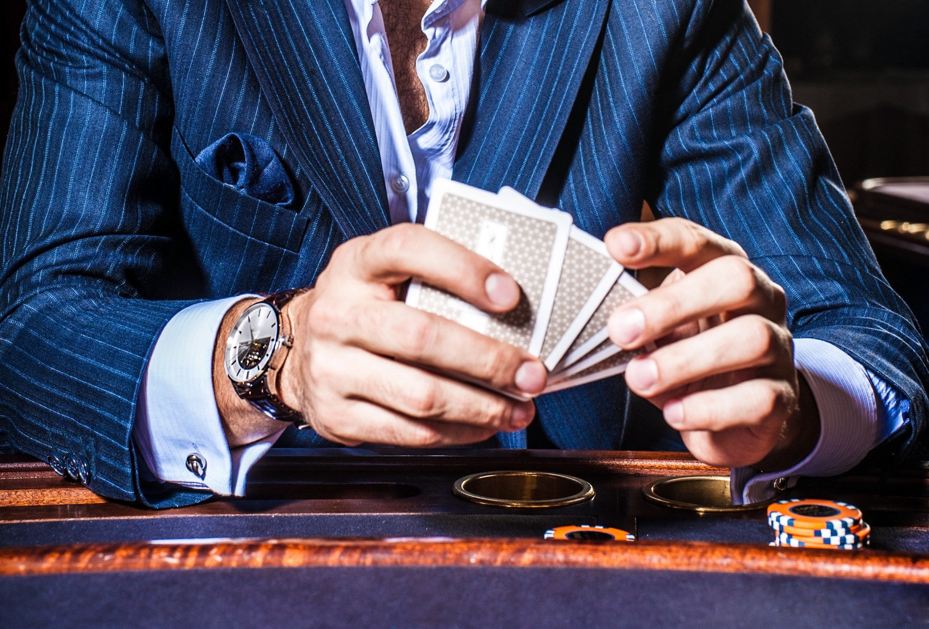 Мужчина в строгом костюме с дорогими часами на руках держит карты, сидя за столом для игры в покер