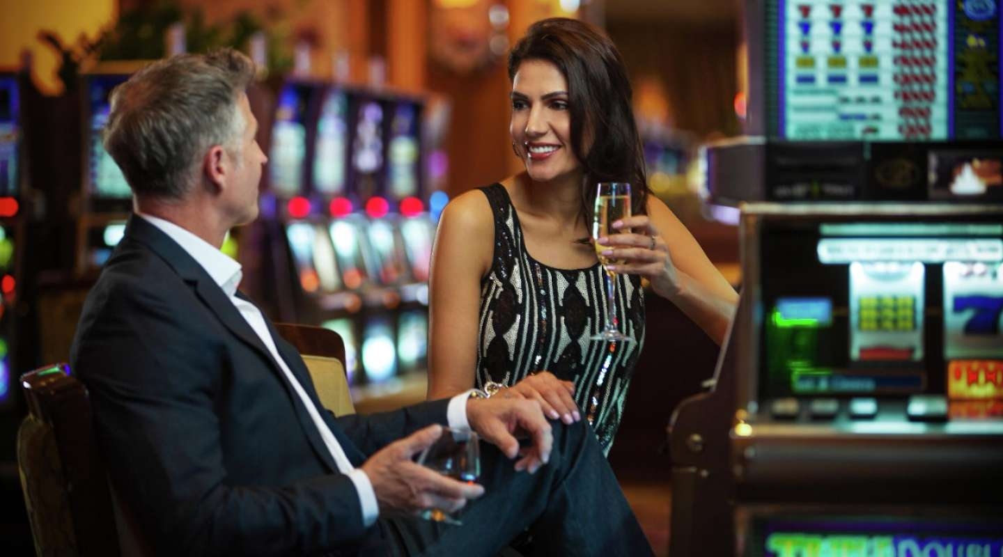 Мужчина играет на игровом автомате, а его спутница, красотка в вечернем платье, пьет шампанское, стоя рядом