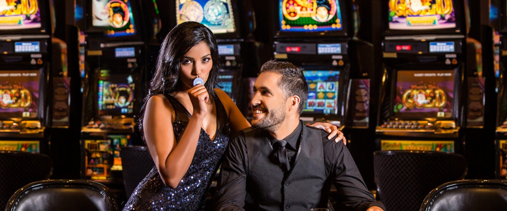 Мужчина с довольной ухмылкой сидит в кресле в зале с игровыми автоматами, а его сексуальная спутница целует фишку