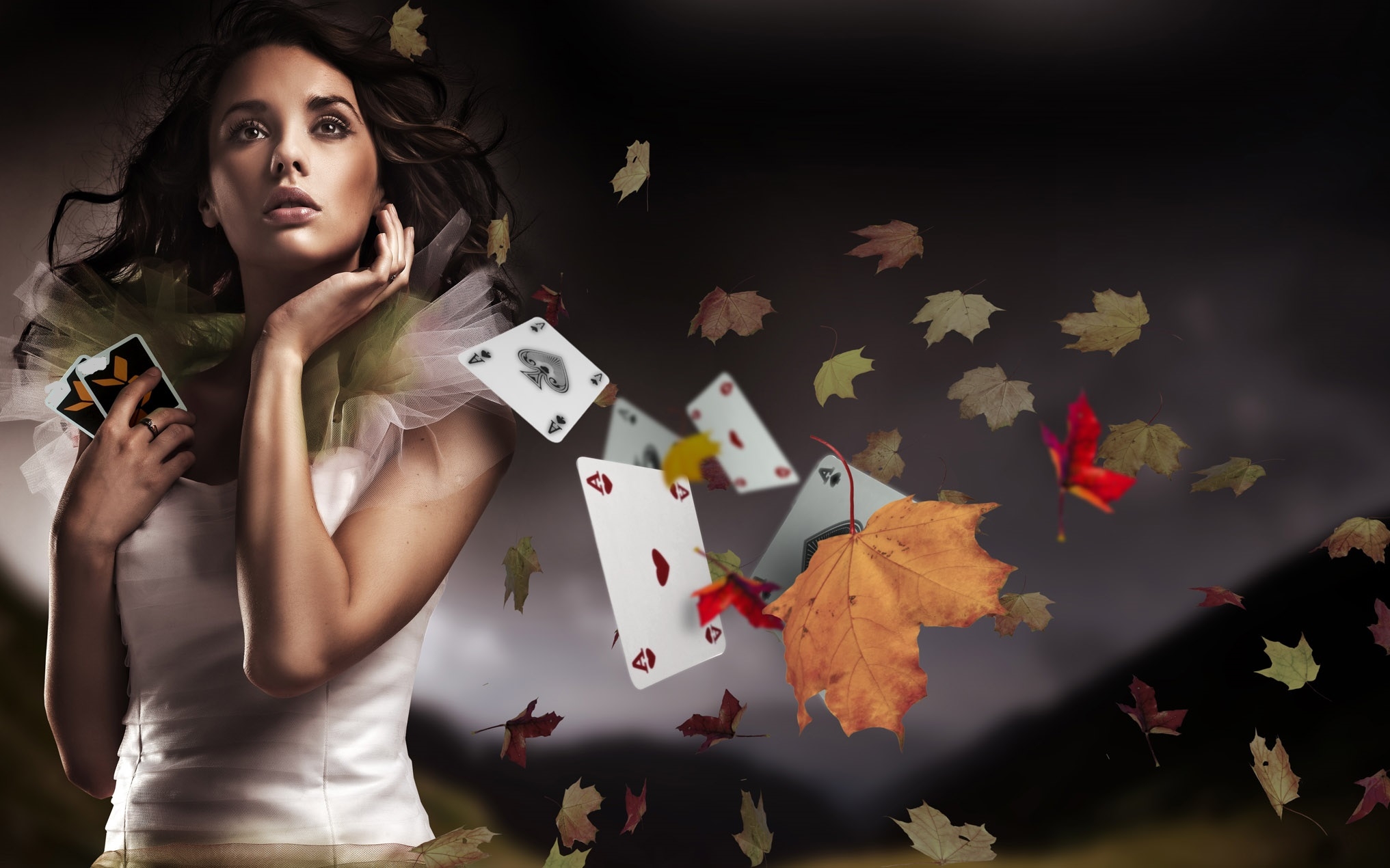 Привлекательная красотка с картами в руках задумчиво смотрит вверх, а рядом пролетают осенние листья и игральные карты