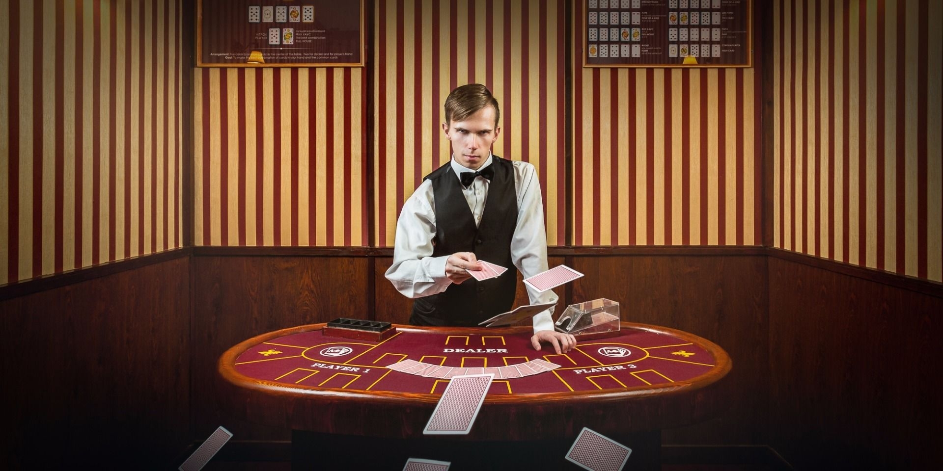 Молодой парень дилер казино одетый в белую рубашку с бабочкой, черную жилетку строгие брюки стоит у игорного стола