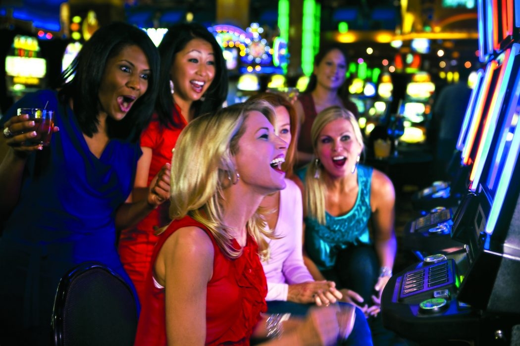 Девушке с европейской внешностью, азиатка, мулатка и еще пару человек на заднем фоне восторженно кричат, направив свои взоры на монитор игрового автомата