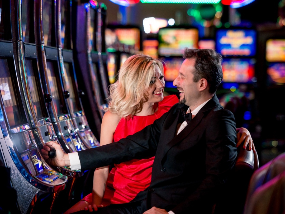 Блондинка в красном платье хорошо проводит время за игрой на автоматах в кампании своего богатого мужчины