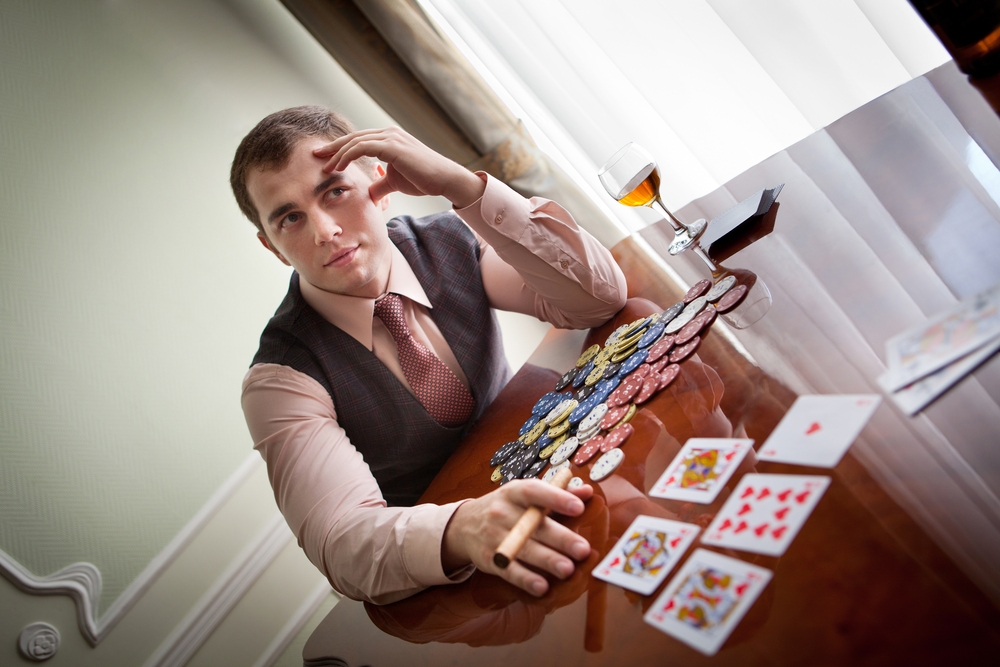 Молодой мужчина в деловой одежде сидит за столом, на котором лежат фишки для покера и стоит бокал с коньяком