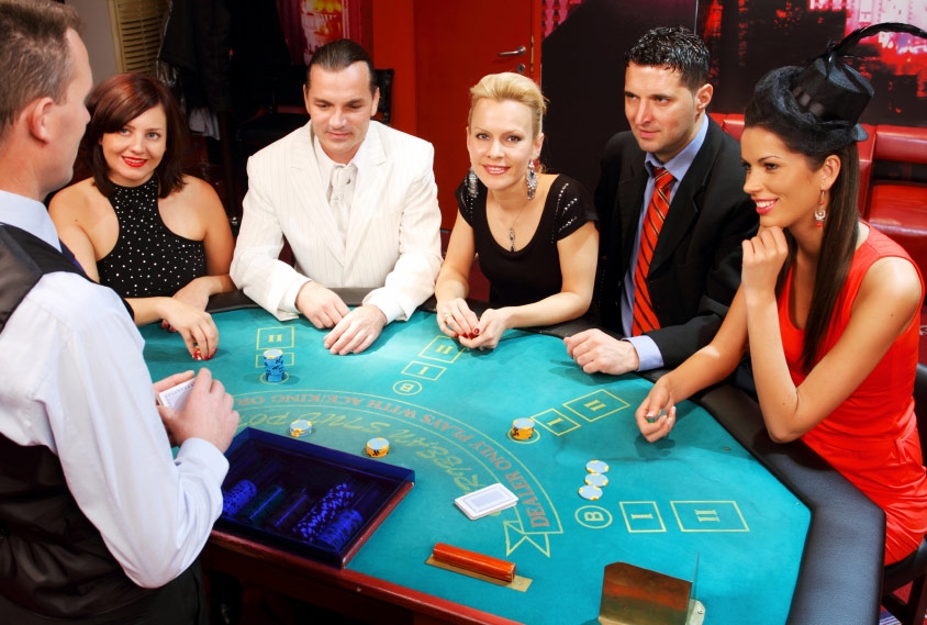 Мужчины  в деловых костюмах и женщины в вечерених платьях собрались за столом для покера, чтобы сыграть партию в компании крупье