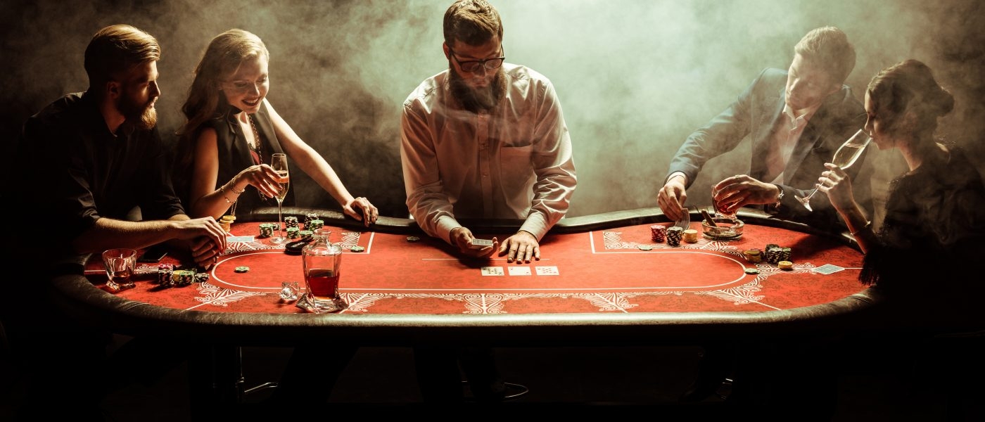 Бородатый молодой мужчина крупье ведет игру в покер для компании богатых парней и девушек, которые распивают шампанское и виски в задымленной комнате за столом для покера