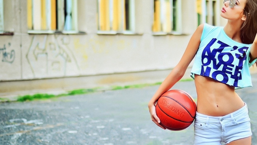 Стройная девушка в мини-шертах и топике позирует на улице с баскетбольным мячом в руках