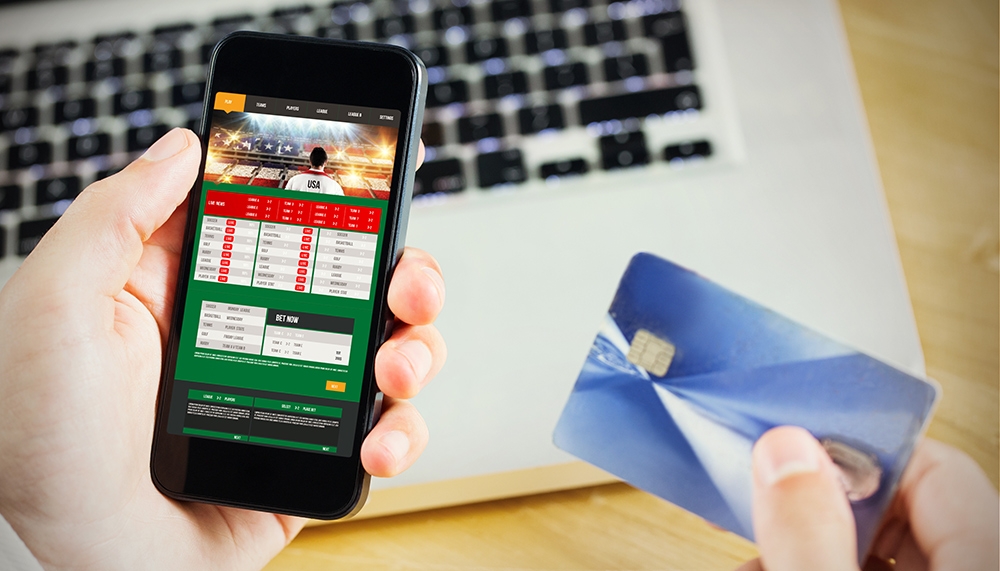 Человек держит в одной руке смартфон с открытым сайтом букмекерской конторы, а во второй банковскую карту