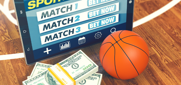 Планшет, на котором открыт сайт букмекерской конторы, миниатюрный баскетбольный мяч и стодолларовые купюры находятся на столе