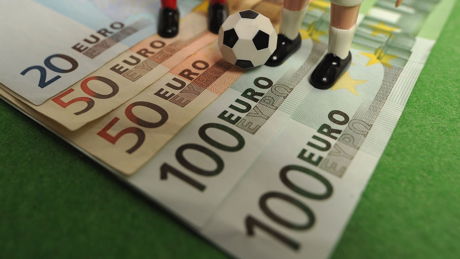 Купюры номиналом 20,50 и 100 евро, на которых стоят игрушечные футболисты и лежит мини-мяч