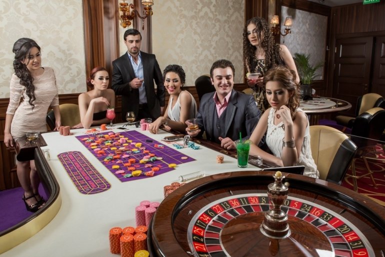Двое мужчин в компании пяти красивых женщин в бежевых коктейльных платьях играют в рулетку в казино вип-уровня
