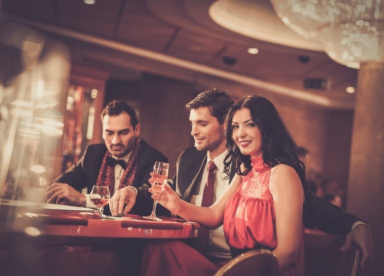 Роскошная брюнетка и двое молодых мужчин в смокингах играют в казино, распивая шампанское