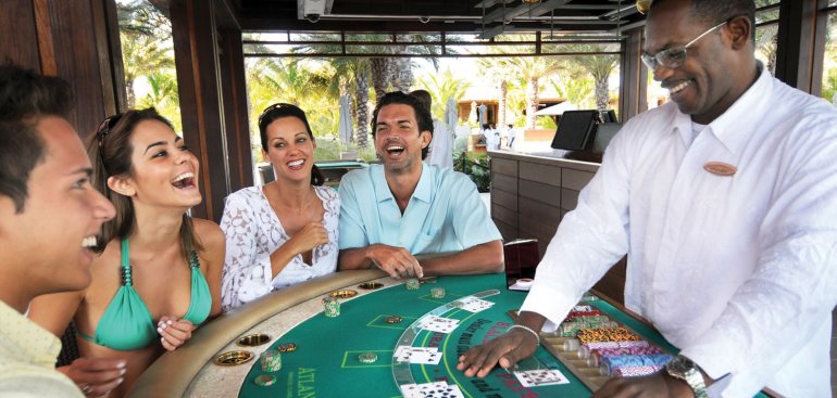 Темнокожий крупье ведет игру в блэкджек для азартных туристов, а женщина в бирюзовом купальнике весело смеется за игорным столом