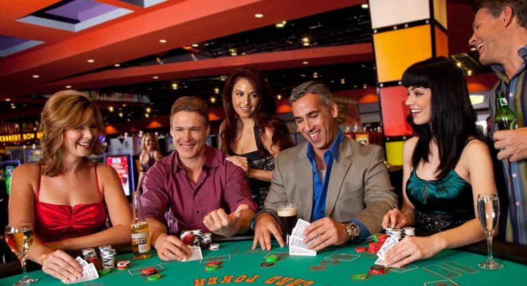 Трое мужчин в костюмах и трое женщин в вечерних платьях смеются и пьют алкоголь за игрой в трехкарточный покер в курортном казино