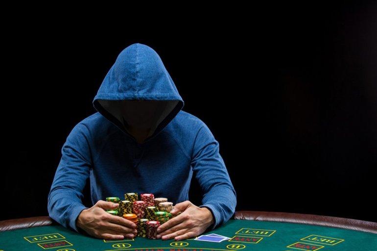 Мужчина скрывает лицо за капюшоном, сидя у стола для покера перед горой фишек