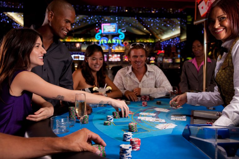 Темнокожий мужчина и его красивая спутница в вечернем платье играют в блєкджек в компании других гостей казино и молодой девушки дилера